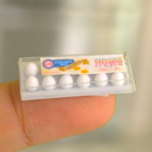 May include: Miniature plastic egg carton containing six white eggs. The carton has a clear plastic top and a label with the words "EGGSAMIN BEST" and "VITAMINS A-D-E-B12". The carton is held by a finger.