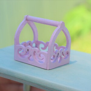 May include: A small, pink, wooden decorative basket with a handle and intricate cut-out designs. The basket is sitting on a light blue surface.