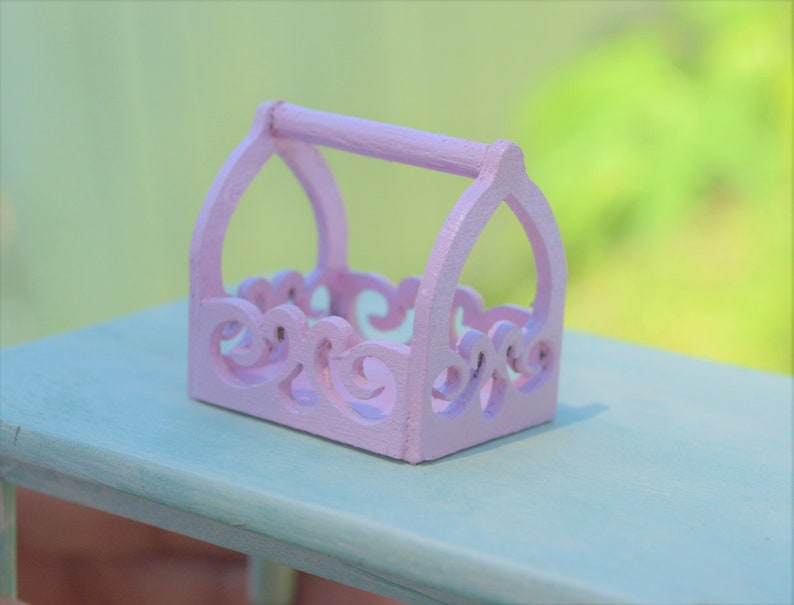 May include: A small, pink, wooden decorative basket with a handle and intricate cut-out designs. The basket is sitting on a light blue surface.