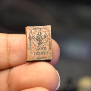 May include: A small, brown book with an intricate cover design. The cover displays a geometric symbol within a triangle and the Greek text "&Omega;&Chi;&Epsilon; &Iota;&Omicron;&Chi;&Upsilon;&Sigma;". The book is held between fingers.