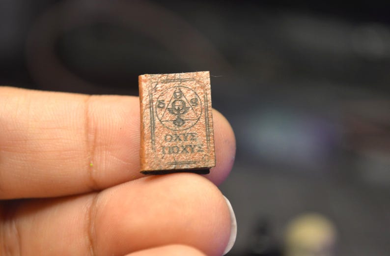 May include: A small, brown book with an intricate cover design. The cover displays a geometric symbol within a triangle and the Greek text "&Omega;&Chi;&Epsilon; &Iota;&Omicron;&Chi;&Upsilon;&Sigma;". The book is held between fingers.