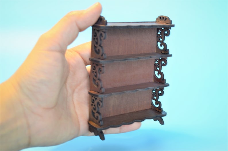 Pu&ograve; includere: Un'unit&agrave; a scaffale in legno in miniatura con tre ripiani e dettagli intricati a volute. Lo scaffale &egrave; dipinto di un colore marrone scuro.