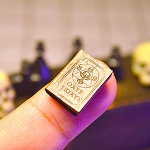 May include: A small, aged book with engraved symbols and text, held between fingers. The book has a light brown cover and dark text. The background includes skulls and bottles, suggesting a mystical or occult theme.