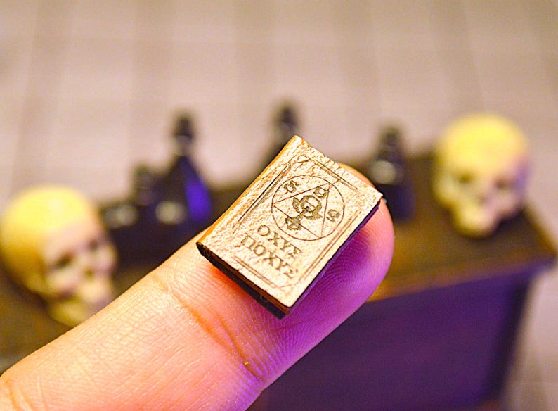 May include: A small, aged book with engraved symbols and text, held between fingers. The book has a light brown cover and dark text. The background includes skulls and bottles, suggesting a mystical or occult theme.