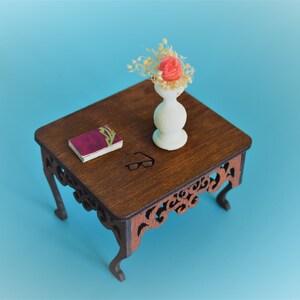 May include: A miniature wooden table with intricate carvings on the legs. A small book and a white vase with a single pink flower and dried foliage are on the table.