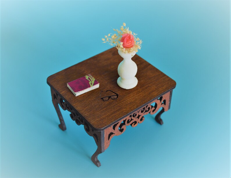 May include: A miniature wooden table with intricate carvings on the legs. A small book and a white vase with a single pink flower and dried foliage are on the table.