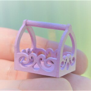 May include: A small, light pink wooden basket with a decorative, ornate design and a handle. The basket is perfect for holding small items.