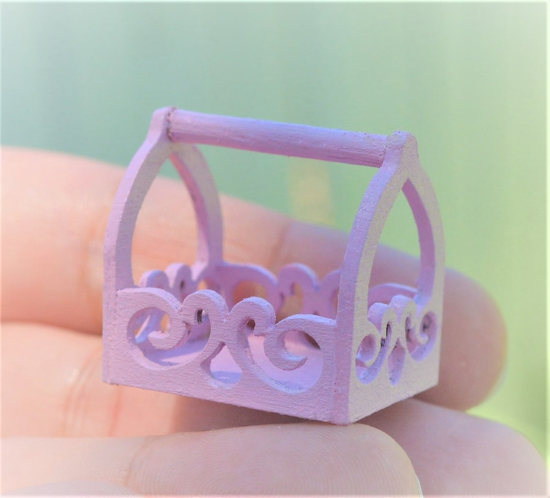 May include: A small, light pink wooden basket with a decorative, ornate design and a handle. The basket is perfect for holding small items.