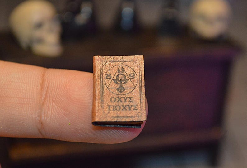 May include: A tiny, aged book with a brown cover. The cover has a black geometric design and text in a foreign language. The book is held between two fingers, indicating its small size. The background is blurred, with suggestions of skulls and dark furniture.