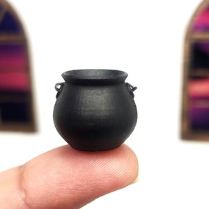 Op de afbeelding: Een kleine zwarte ketel met twee handvatten. De ketel staat op een vinger.