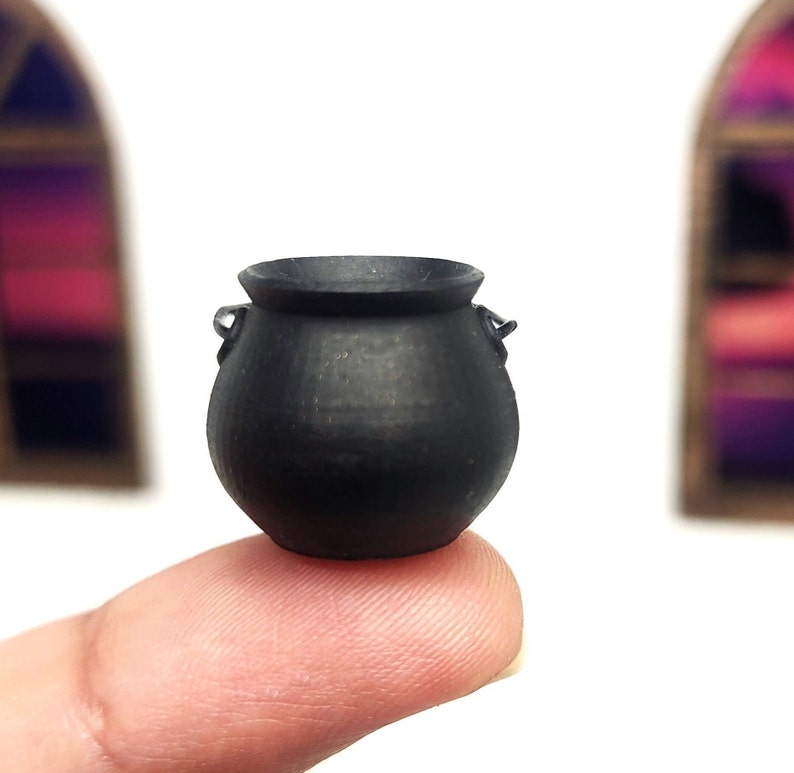 Op de afbeelding: Een kleine zwarte ketel met twee handvatten. De ketel staat op een vinger.