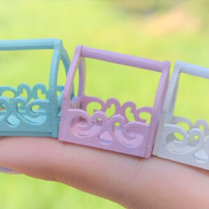 May include: Three miniature decorative baskets in pastel colors. The baskets are made of wood and have a cutout design. The baskets are painted in light blue, pink, and white.
