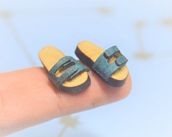 May include: A pair of miniature teal leather sandals with two straps and a brown sole.