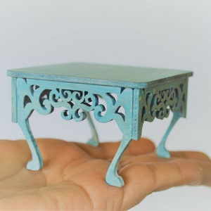 May include: A small, light blue, wooden table with intricate cutouts and decorative legs. The table is about 6 inches wide and 4 inches tall.