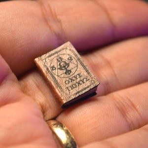 May include: A tiny book with a brown cover and black pages. The cover has a circular design with symbols and text. The book is held in a hand, indicating a small size, perhaps for a dollhouse or as a collectible item.