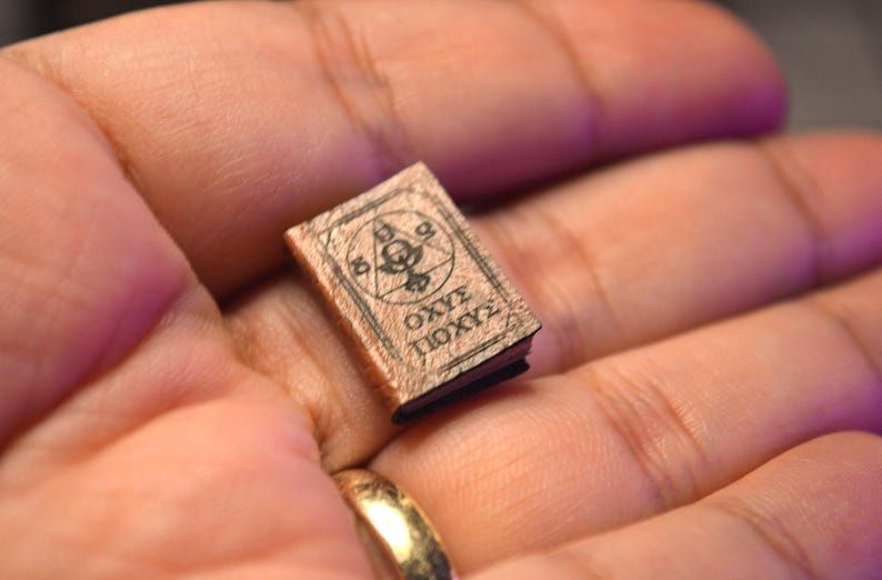 May include: A tiny book with a brown cover and black pages. The cover has a circular design with symbols and text. The book is held in a hand, indicating a small size, perhaps for a dollhouse or as a collectible item.