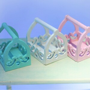 May include: Three decorative wooden baskets with intricate cutouts. The baskets are painted in pastel colors: light blue, white, and pink. The baskets have handles and are arranged in a row on a light green table.