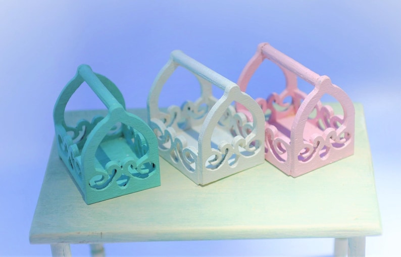 May include: Three decorative wooden baskets with intricate cutouts. The baskets are painted in pastel colors: light blue, white, and pink. The baskets have handles and are arranged in a row on a light green table.
