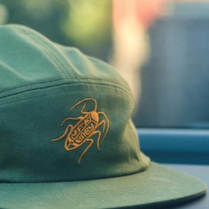 Puede incluir: Gorra verde de cinco paneles con visera plana. La gorra presenta un diseño de insecto naranja bordado con el texto "MFK BABY". El sombrero está hecho de una tela duradera y tiene un estilo clásico e informal.