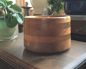 Puede incluir: Un tazón de madera con un acabado natural y un diseño en capas. El tazón está hecho de varias piezas de madera, creando un aspecto único y rústico.