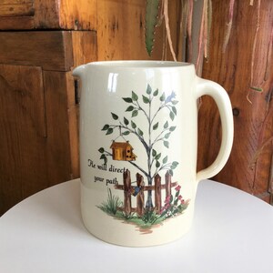 Puede incluir: Una jarra de cerámica de color crema con un diseño pintado a mano de un árbol con una casita para pájaros, una valla y flores. La jarra tiene una cita que dice "Él dirigirá tu camino".