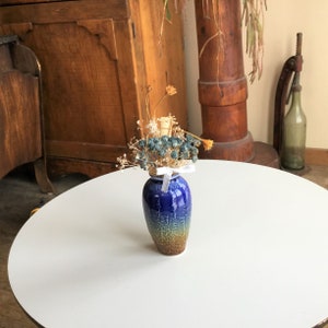 Op de afbeelding: Een kleine, blauwe keramische vaas met een textuurpatroon en een verloop van blauw naar bruin. De vaas is gevuld met gedroogde bloemen en groen, vastgebonden met een wit lint. De vaas staat op een witte tafel.