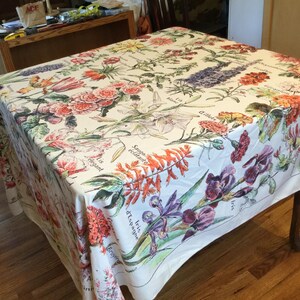 May include: A rectangular tablecloth with a white background, adorned with a colorful floral pattern. The design features various flowers in shades of red, pink, purple, orange, and green, with botanical labels in French. The tablecloth is draped over a wooden table.