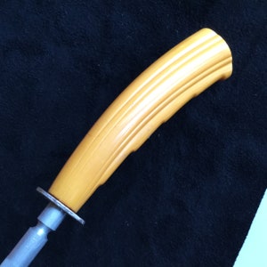 Op de afbeelding: Een vintage messenslijper met een lichtgele, geribbelde handgreep. De handgreep is gebogen en bevestigd aan een zilverkleurige metalen staaf en een cirkelvormige bescherming. De slijper is ontworpen voor gebruik in de keuken.