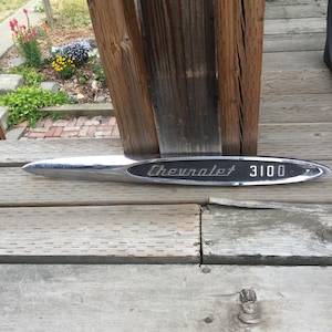 May include: A vintage Chevrolet 3100 emblem, featuring a chrome finish and a black oval center. The text "Chevrolet" and "3100" are displayed in a classic font. The emblem is elongated with a pointed end.