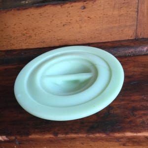 Puede incluir: Una tapa de vidrio ovalada de color verde pálido con un asa elevada. La tapa tiene un acabado liso y brillante y está colocada sobre una superficie de madera. El asa es de un tono verde más claro.