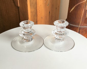 May include: Two clear glass candle holders with a tiered design. Each holder has a round base and a central pillar with a cup for a candle.