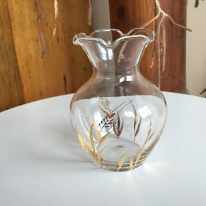 Puede incluir: Un jarrón de cristal transparente con borde ondulado y cuerpo redondeado. El jarrón presenta un diseño de espigas de trigo doradas. El jarrón está sobre una superficie blanca.