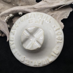 May include: A round, off-white ceramic lid featuring a central cross-shaped knob. Embossed text around the perimeter reads THE VEIR No 2 PAT MAR 1 02 APRIL 16 1901. The surface has a smooth, glazed finish with visible raised lettering.
