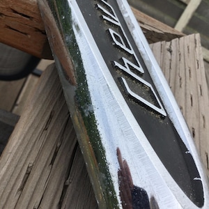 May include: Close-up of a chrome and black metal sign with the word "Chevy" in white lettering. The sign is angled, showing its reflective surface and the wooden background.