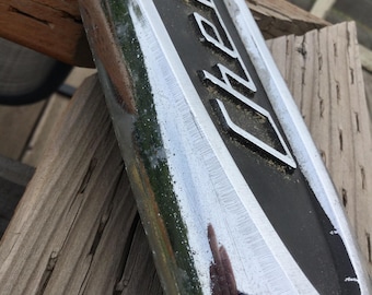 Puede incluir: Primer plano de un letrero de metal cromado y negro con la palabra "Chevy" en letras blancas. El letrero está en ángulo, mostrando su superficie reflectante y el fondo de madera.