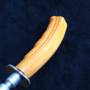 Op de afbeelding: Een vintage meshandvat met een gebogen, amberkleurige greep. De handgreep heeft een geribbeld ontwerp en is bevestigd aan een zilverkleurige metalen bolster en tang. De meshandgreep staat tegen een donkere achtergrond.
