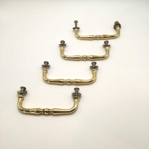 May include: Four polished brass drawer pulls arranged in a diagonal line. Each handle features a smooth, curved U-shape with a central decorative bead detail and two metal mounting screws with washers attached at the ends of the handle arms.