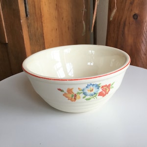 May include: Cream-colored ceramic bowl with a red rim and floral design. The bowl features a pattern of blue, orange, and red flowers. The bowl has a ribbed exterior and is likely vintage or retro.