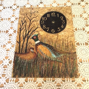 Puede incluir: Un reloj de pared rústico con una escena de faisanes pintada a mano. La obra de arte representa dos faisanes en un campo con hierba alta y árboles desnudos, sobre un fondo dorado texturizado. La esfera del reloj es negra con números dorados.