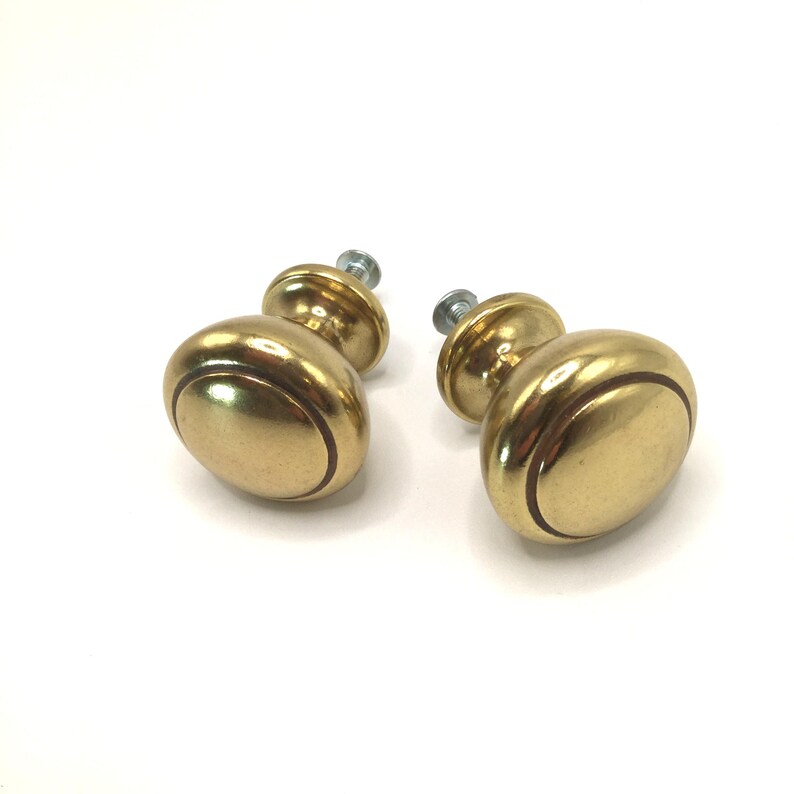 May include: Two round cabinet knobs made of polished brass with a smooth, reflective surface. Each knob features a circular indentation near the edge and includes a silver-colored metal mounting screw protruding from the back of the base.