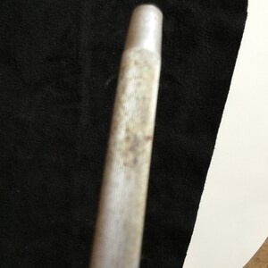 Op de afbeelding: Close-up van een metalen gereedschap met een cilindrische vorm. Het gereedschap heeft een getextureerd, geribbeld oppervlak met wat verkleuring. Het uiteinde van het gereedschap is een lichter, gladder metaal. De achtergrond is een donkere stof en een wit oppervlak.