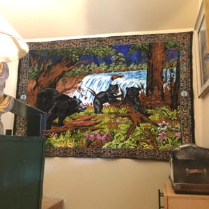 Pode incluir: Uma tapeçaria que retrata uma cena de floresta com uma cachoeira e três ursos negros. Os ursos estão em primeiro plano, com a cachoeira e as árvores ao fundo. A tapeçaria tem uma borda decorativa.