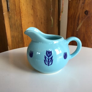May include: A light blue ceramic creamer with a blue floral design. The creamer has a rounded body and a handle.