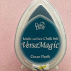 May include: A teal VersaMagic multi-surface chalk ink pad in an egg-shaped, clear plastic case. The ink is labeled "Ocean Depth". The case has a white rim and the brand name "Dove Drop" is visible.