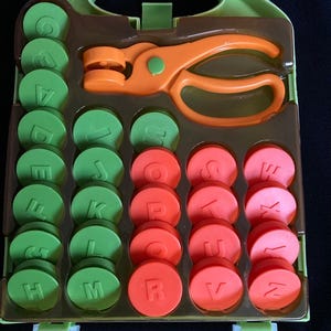 Puede incluir: Un juego de letras del alfabeto verde y naranja con un estuche. El juego incluye fichas de letras circulares verdes y rojas, y una herramienta naranja en forma de tijera. Las letras son visibles en las fichas verdes.