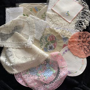 May include: A collection of assorted vintage-style doilies and napkins featuring various textures, including white lace, intricate crochet patterns, and embroidered floral motifs in shades of pink, blue, and green, layered together in a group.