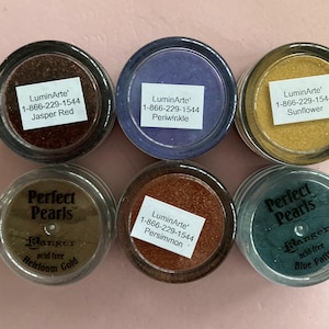 Könnte beinhalten: Sechs Gläser mit Bastelpigmentpulvern in verschiedenen Farben, darunter Rot, Lila, Gold und Grün. Die Gläser haben Etiketten mit Produktnamen und einer Telefonnummer. Die Gläser sind mit "Jasper Red", "Periwinkle", "Sunflower", "Heirloom Gold", "Persimmon" und "Blue Patina" beschriftet.