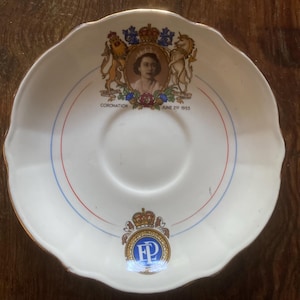 Op de afbeelding: Witte schotel met een geschulpte rand en gouden rand. De schotel heeft een portret, de tekst "CORONATION JUNE 2nd 1953" en een wapen met een kroon en de initialen "ER". Rode en blauwe concentrische cirkels omcirkelen het midden.