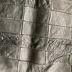 May include: Close-up of a black leather material with a patchwork design. The leather has a textured surface with visible stitching, creating a grid-like pattern. The colour is a deep, solid black.