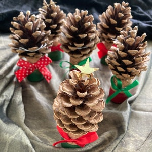 May include: A selection of handmade Christmas decorations. Each ornament is made from a pine cone fixed to a cork base, decorated with colourful ribbons and a star-shaped embellishment. They are arranged on a textured, grey surface.